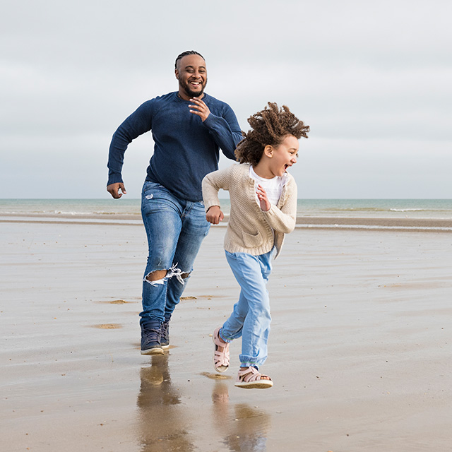 A young black girl wearing jeans and a tan shirt laughs and runs in front of her father on a sandy beach