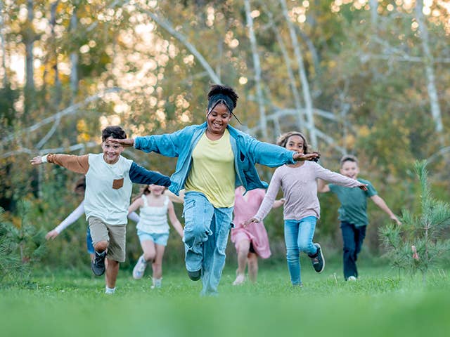 Group of boys and girls run through a grassy field outdoors with their arms stretched out to their sides