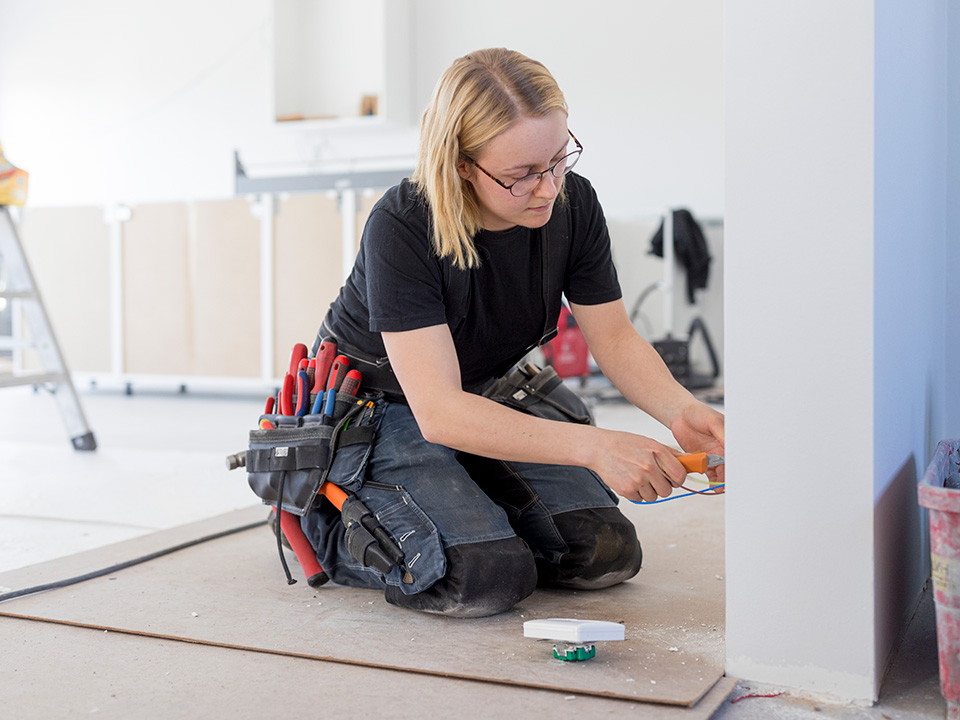 Middle aged white woman rests on her knees as she does electrical work on a construction site