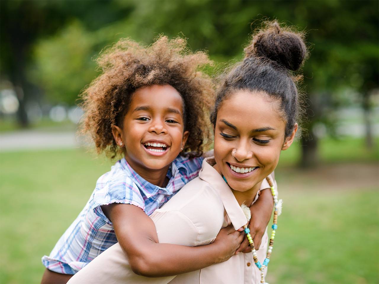 Smiling Black woman looks back at her smiling daughter while carrying her on her back outdoors