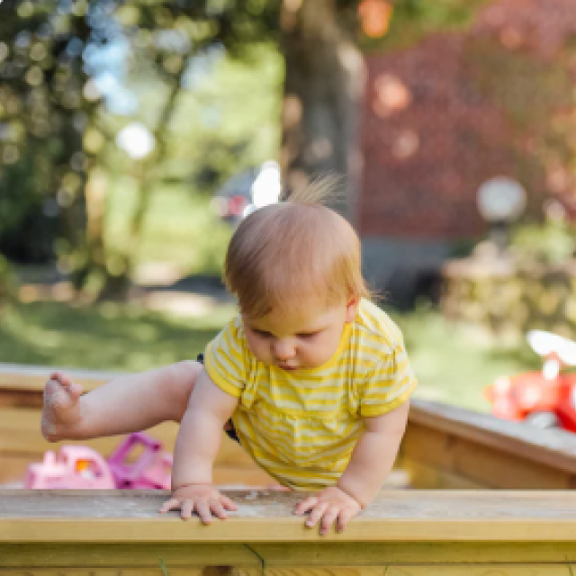 Baby in yellow onesie climbs inside sandbox on sunny day