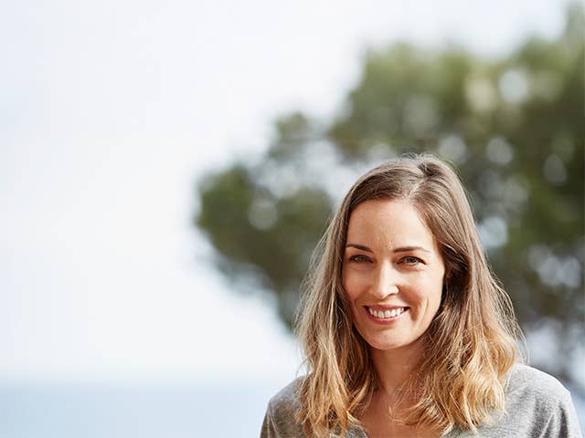 Smiling White woman in a grey shirt stands outdoors in front of a tree