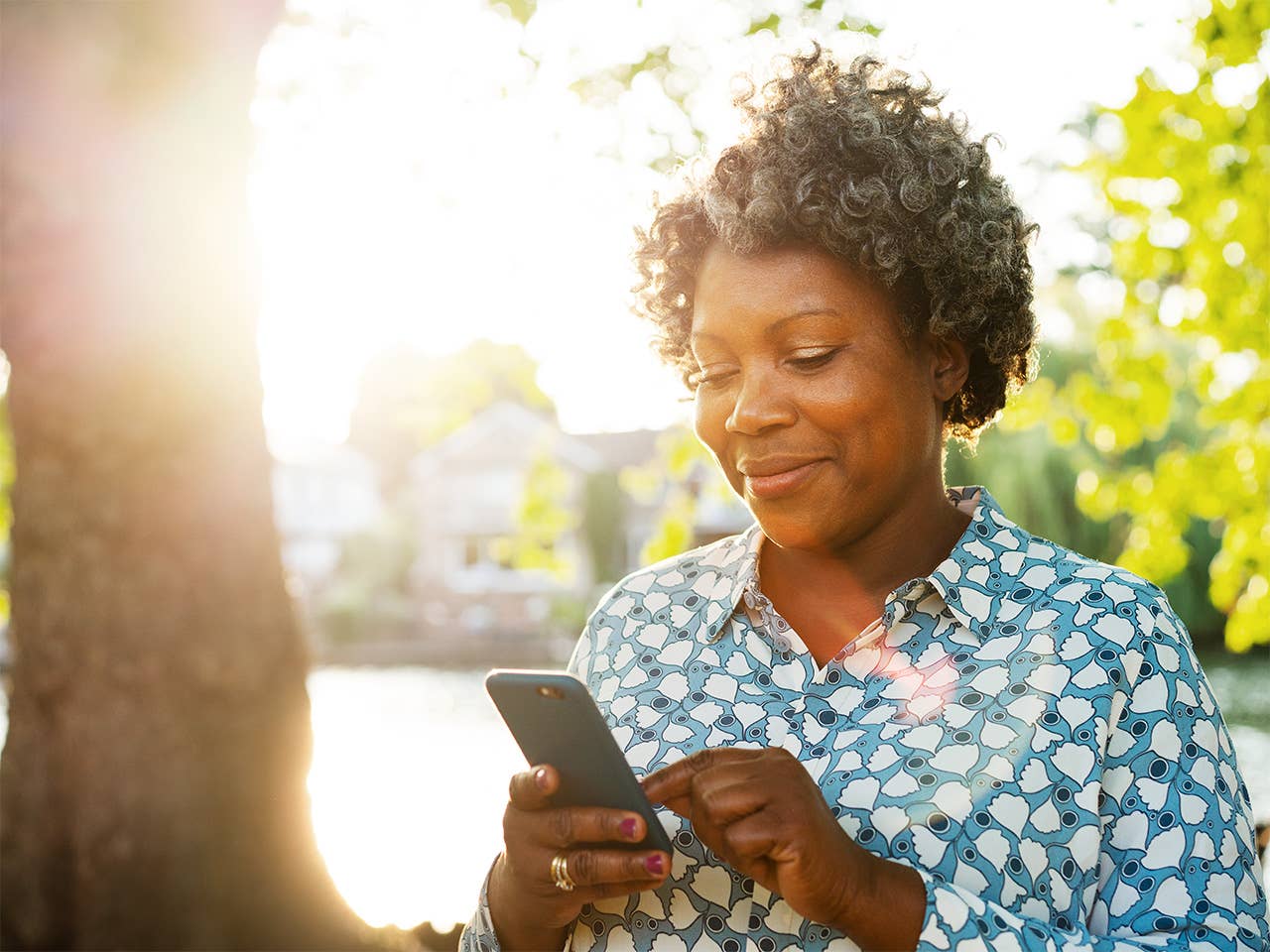 Black woman in a light blue shirt stands beneath a tree in the sunlight, looking at her phone