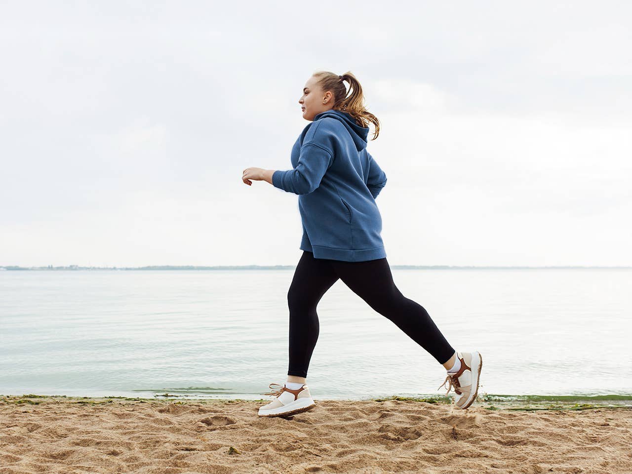 Middle aged white woman in long pants and a sweatshirt jogs alongside a lake