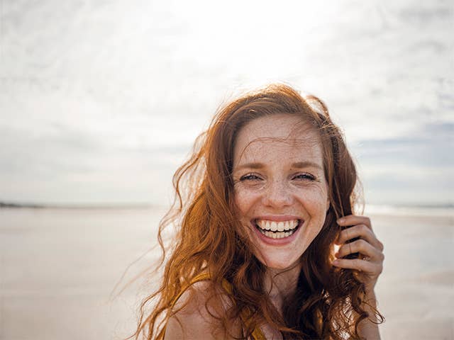 Smiling White woman with red hair and freckles stands outdoors along the beach
