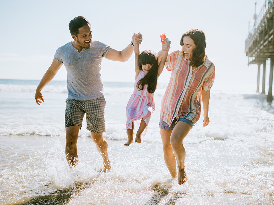 Smiling mother and father lift up young daughter alongside water at the beach