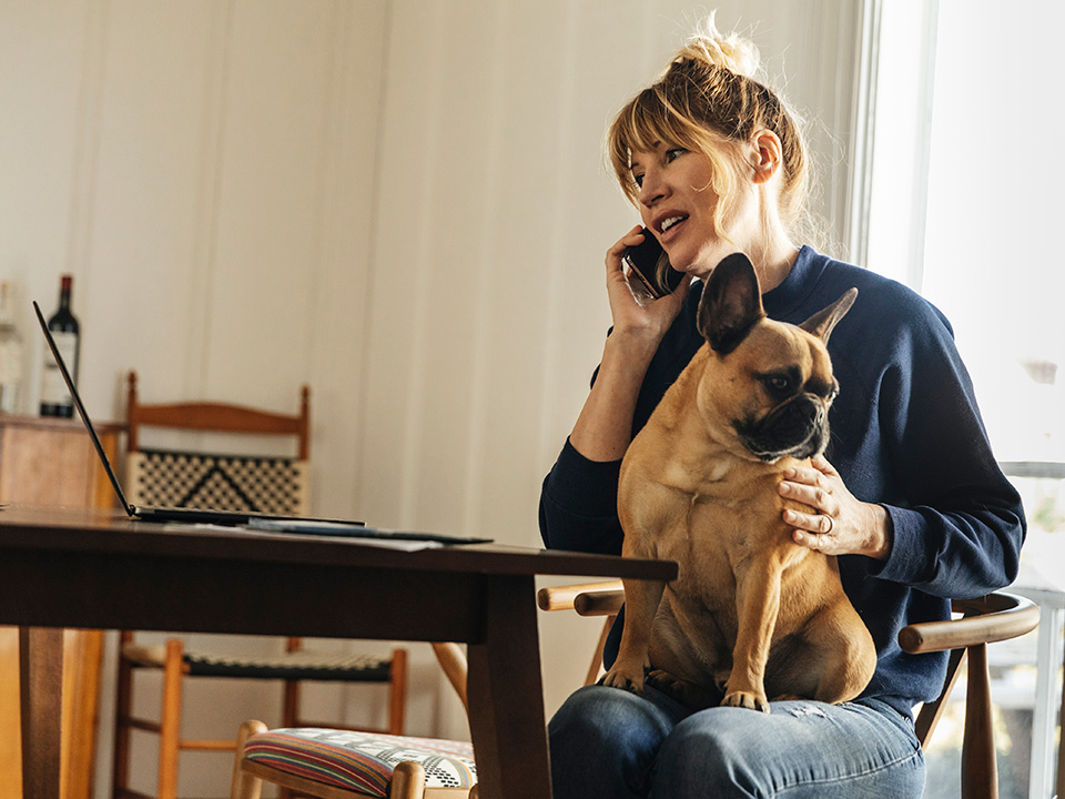 women holding her dog is sitting at the kitchen table scheduling a doctors appointment on her cell phone