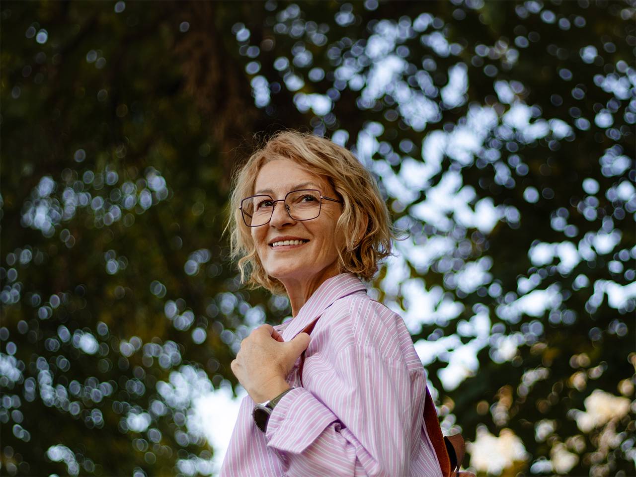 Smiling older white woman wearing glasses looks over her shoulder while in the shade of a tree outdoors