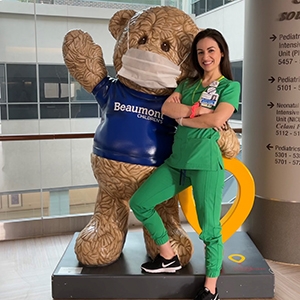 A nurse in green scrubs stands beside a large teddy bear, symbolizing care and comfort in a pediatric setting.