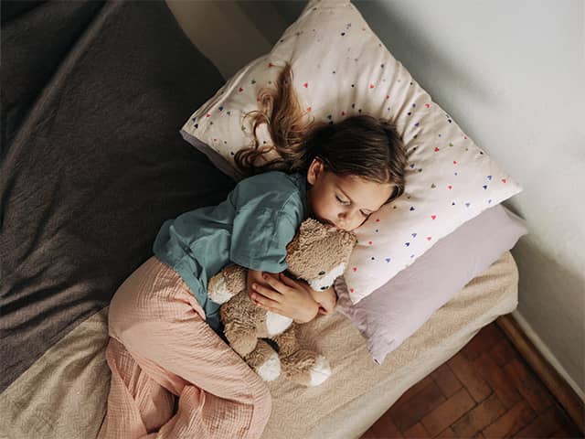 Young girl wearing a blue pajama shirt and white pajama pants sleeps in a bed hugging a teddy bear