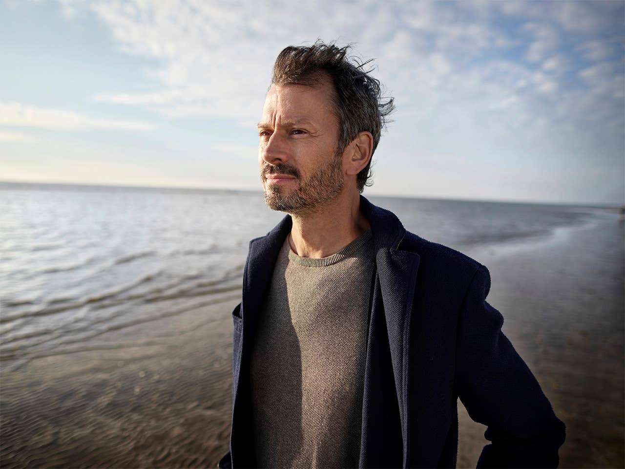 Middle aged white man in a grey zip-up sweatshirt stands alongside a lake