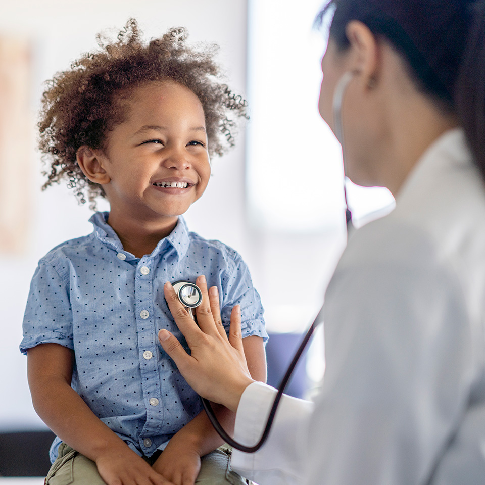 Young boy with curly brown hair smiles as a doctor puts a stethoscope on his chest