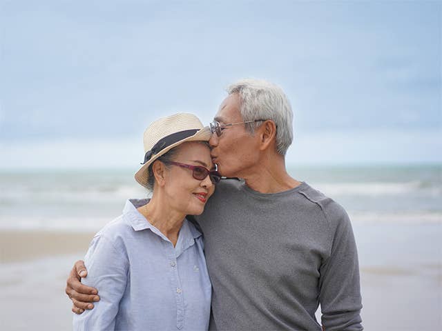 Older Asian man with grey hair and glasses hugs and kisses his wife