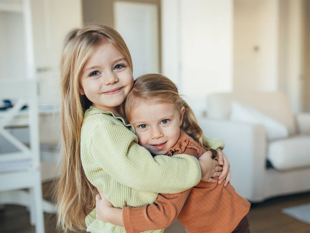 Two young girls with long blonde hair smile and hug each other in a living room