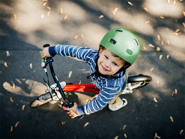 Smiling young boy wearing a blue and white striped shirt and green helmet sits on a bicycle outdoors