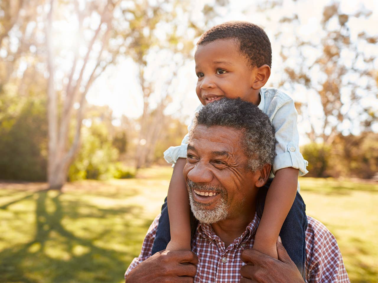 Elderly Black man carries his grandson on his shoulders outdoors in the sunshine