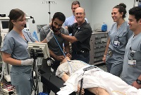 Healthcare workers in scrubs gather around a man in a hospital bed, engaged in a simulation activity.