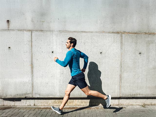 Young white man in black shorts and a blue shirt jogs on the sidewalk in sunshine