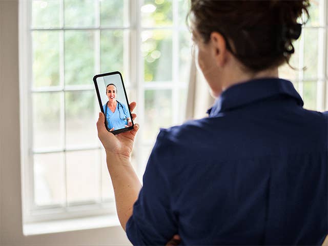 White woman with grey hair sits on her couch holding her phone and speaking with a virtual primary care provider