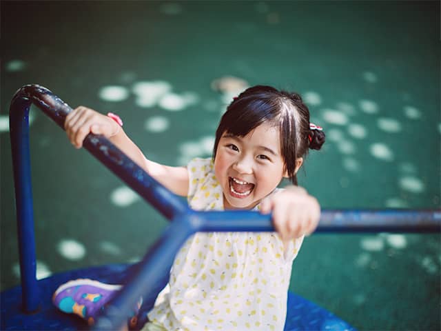 Girl with black hair and wearing a white shirt with yellow polkadots smiles and laughs while riding on a merry go round