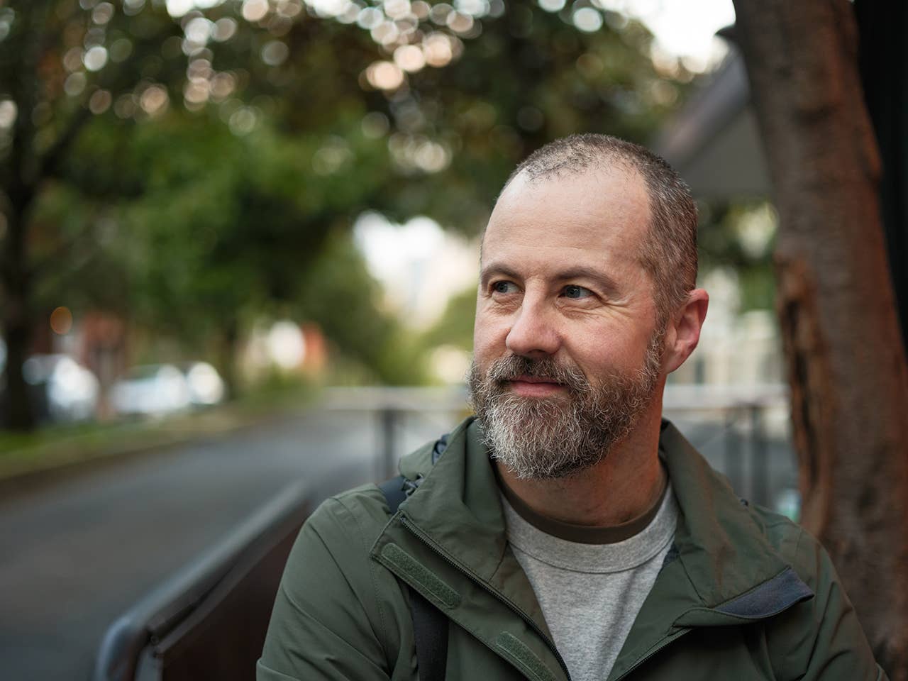 Older white male with balding grey hair and a beard stands outdoors as the evening sun drops below nearby trees