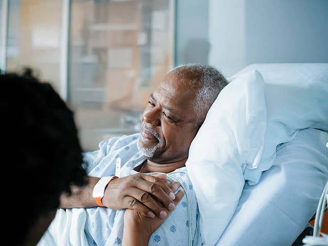 Older Black man laying in a hospital bed smiles and holds hands with his wife