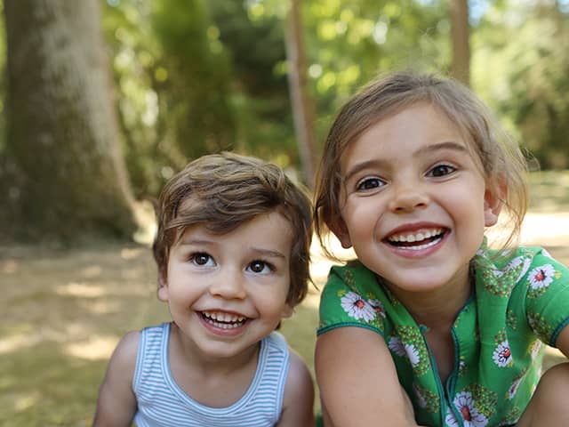 Two children sitting close together outdoors in a wooded park setting, facing the camera.