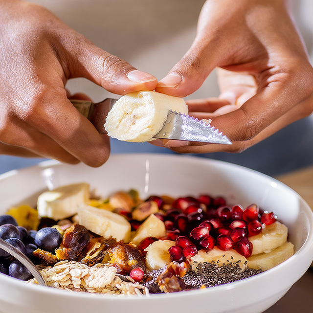 Hands slicing banana over a breakfast bowl with berries, oats, chia seeds and pomegranate seeds on a kitchen counter.