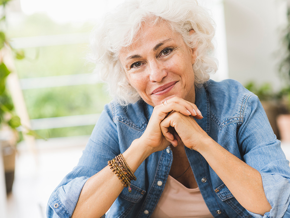 Smiling elderly woman sits and rests her chin on her folded hands