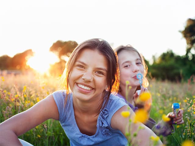 Smiling girl with brown hair and a blue shirt sits beside her sister outdoors in the sunshine