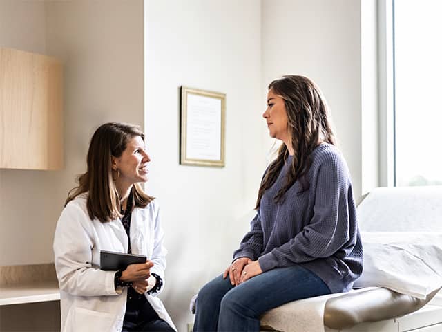 Two women talk in a doctor's office, one seated while discussing health-related topics with the other.