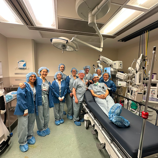 Group of medical trainees and clinicians posed beside an operating table in a surgical suite.