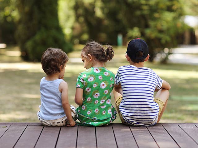 Three children sitting side-by-side on a deck in the shade outdoors