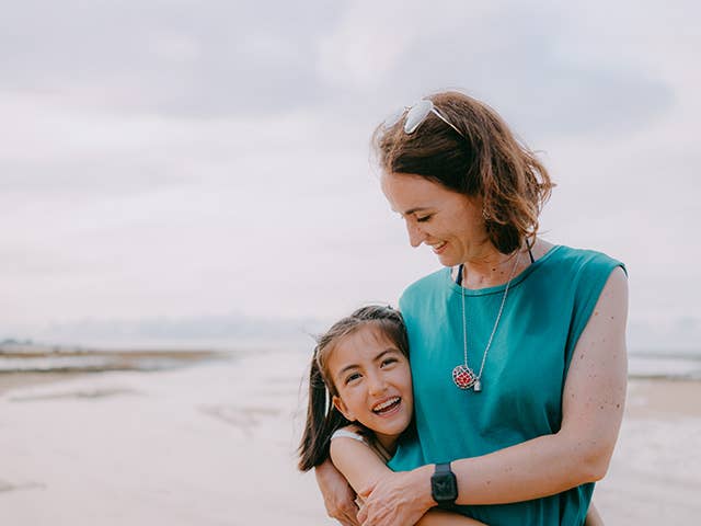 Smiling mother hugs her smiling daughter as they stand beside one another in the sunshine on a sandy beach
