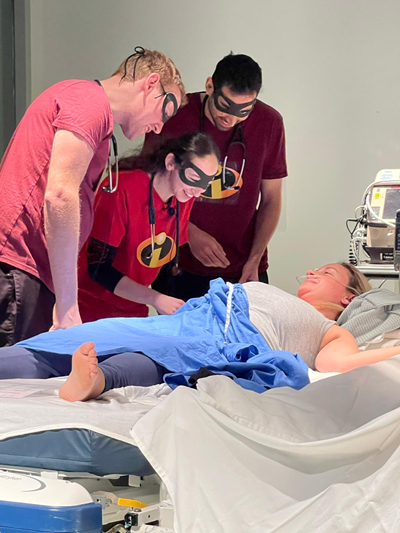 A group of individuals in a hospital room collaborates using a laptop, engaged in pediatric emergency simulation training.