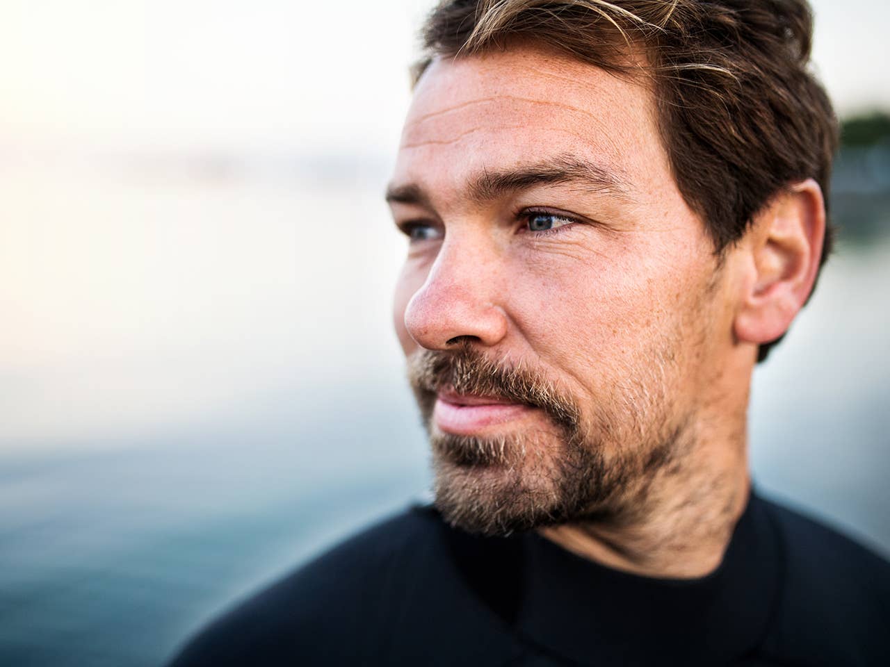 Man with brown hair and a goatee stands next to a lake, looking into the distance