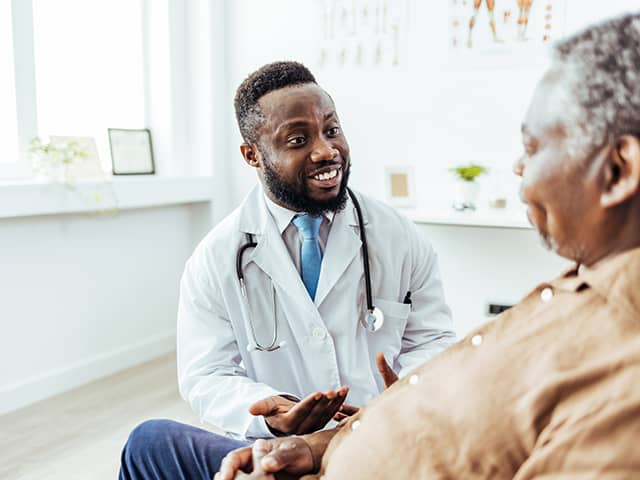 A male doctor converses with an older man in a medical office setting, discussing health concerns.