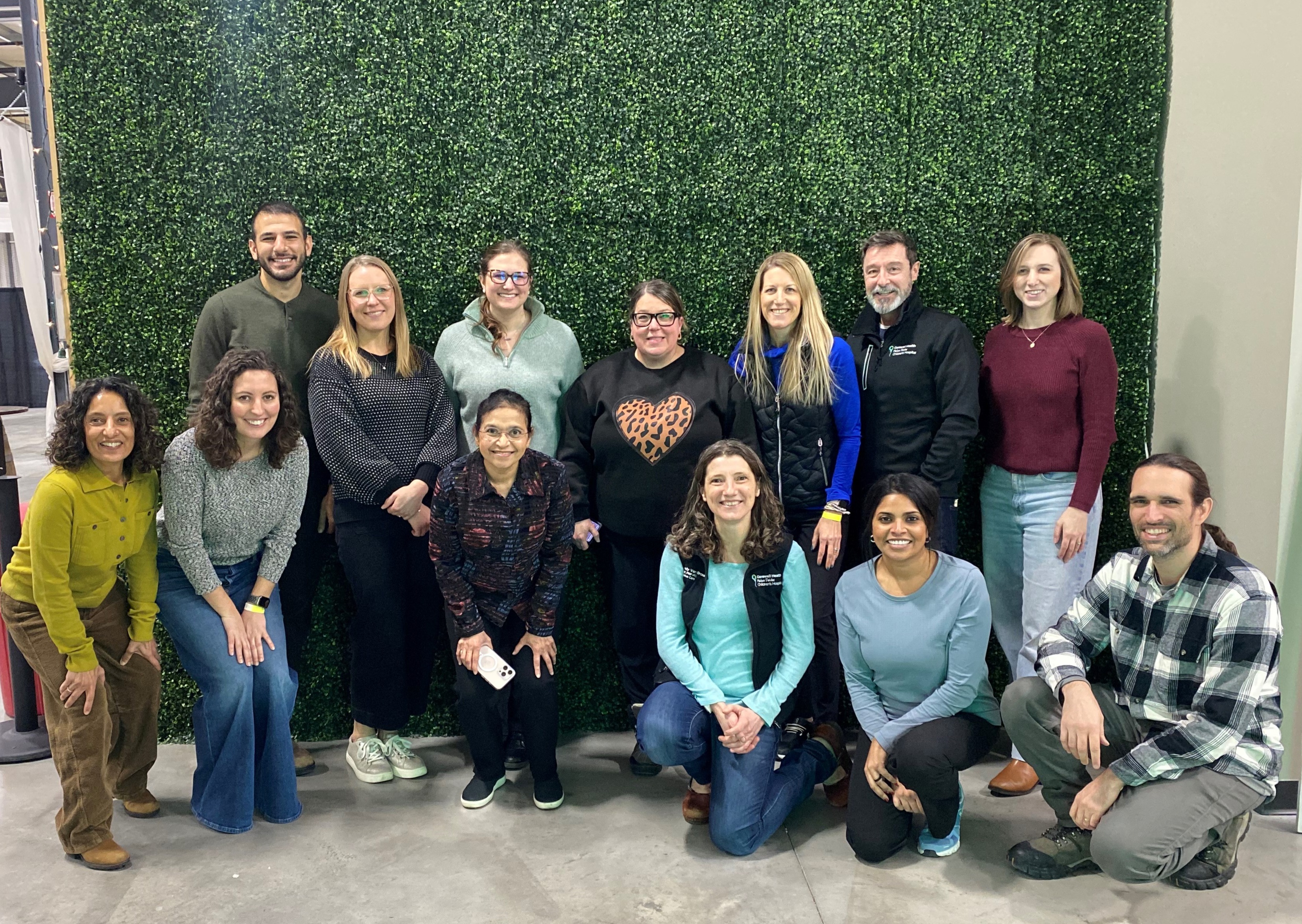 Group of 14 people posing in two rows in front of a green hedge wall indoors.