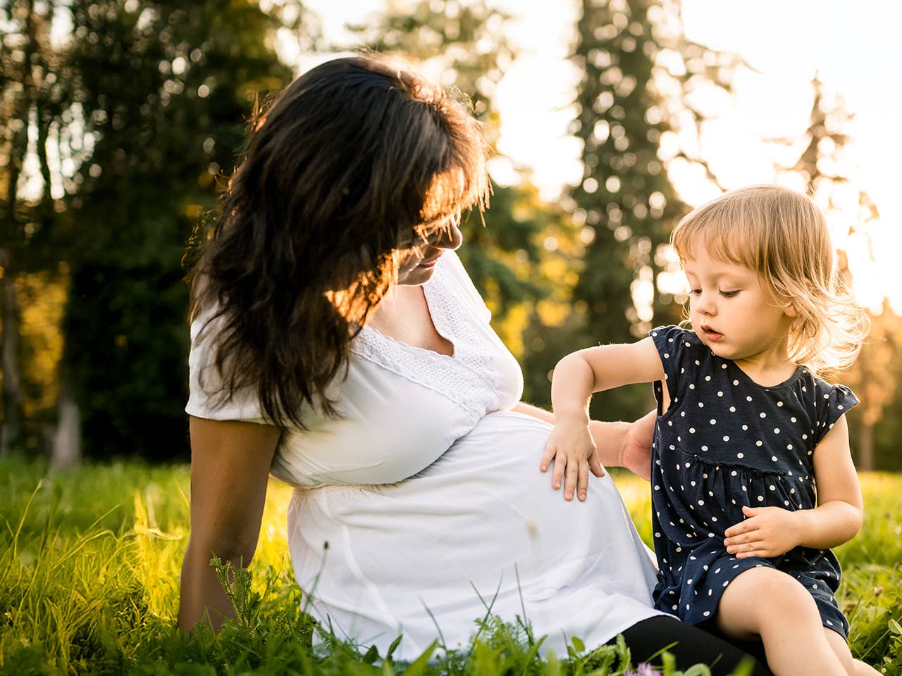 Laughing mother wearing a white dress plays with her toddler in the evening sun