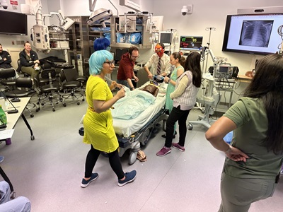 A hospital room scene featuring a patient in bed surrounded by medical staff and fellow trainees engaged in simulation training.