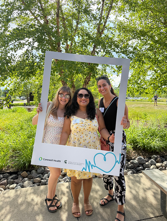 Three individuals standing outdoors on a sunny day, holding a large white photo frame with Corewell Health branding and a heart graphic, with green trees and landscaping in the background.