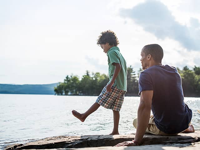 Young Black boy with curly brown hair stands on a large rock near a lake while his father sits next to him