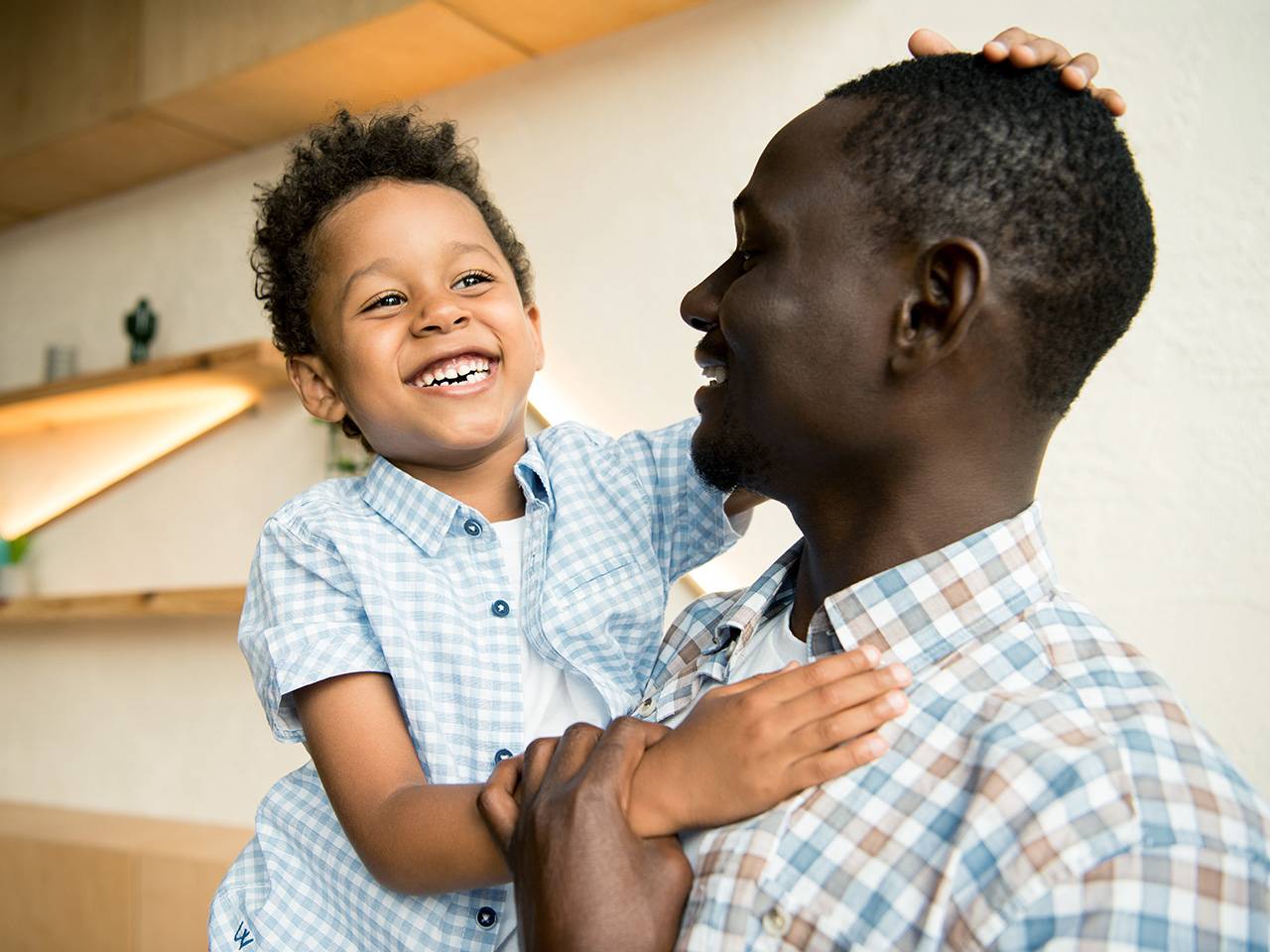 Smiling Black man lifts his smiling son up as they stand inside a sunlit room