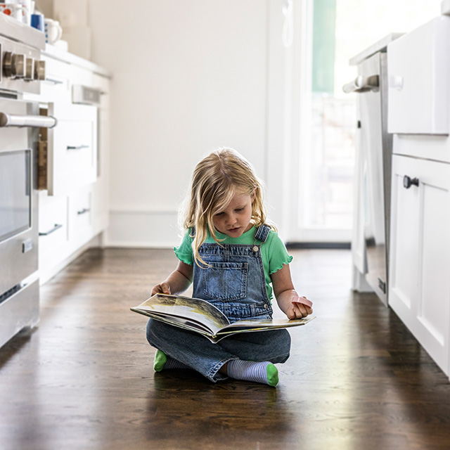 A young girl wearing a green shirt and denim overall sits on the kitchen floor looking at a book
