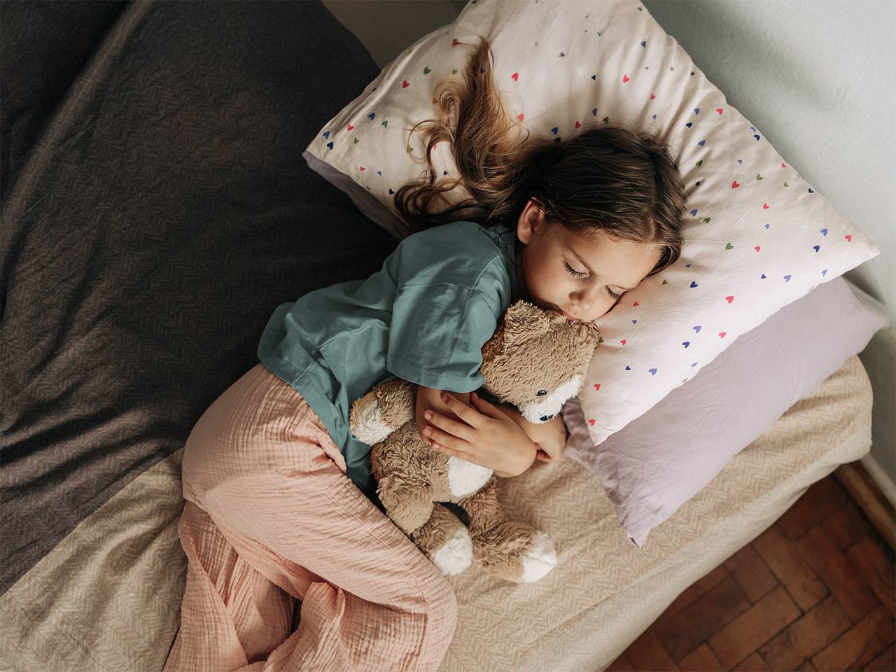 Young girl wearing a blue pajama shirt and white pajama pants sleeps in a bed hugging a teddy bear