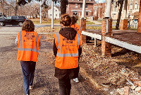 Three individuals in orange vests walking along a street, promoting community engagement and CPR training initiatives.