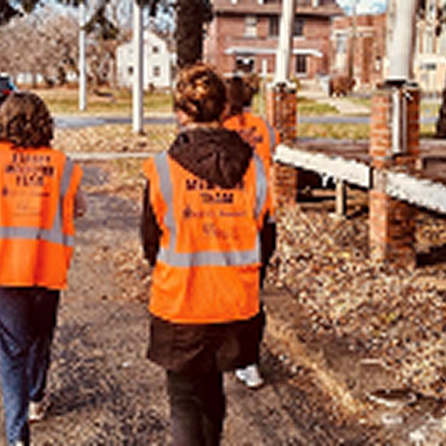 Three individuals in orange vests walking along a street, promoting community engagement and CPR training initiatives.