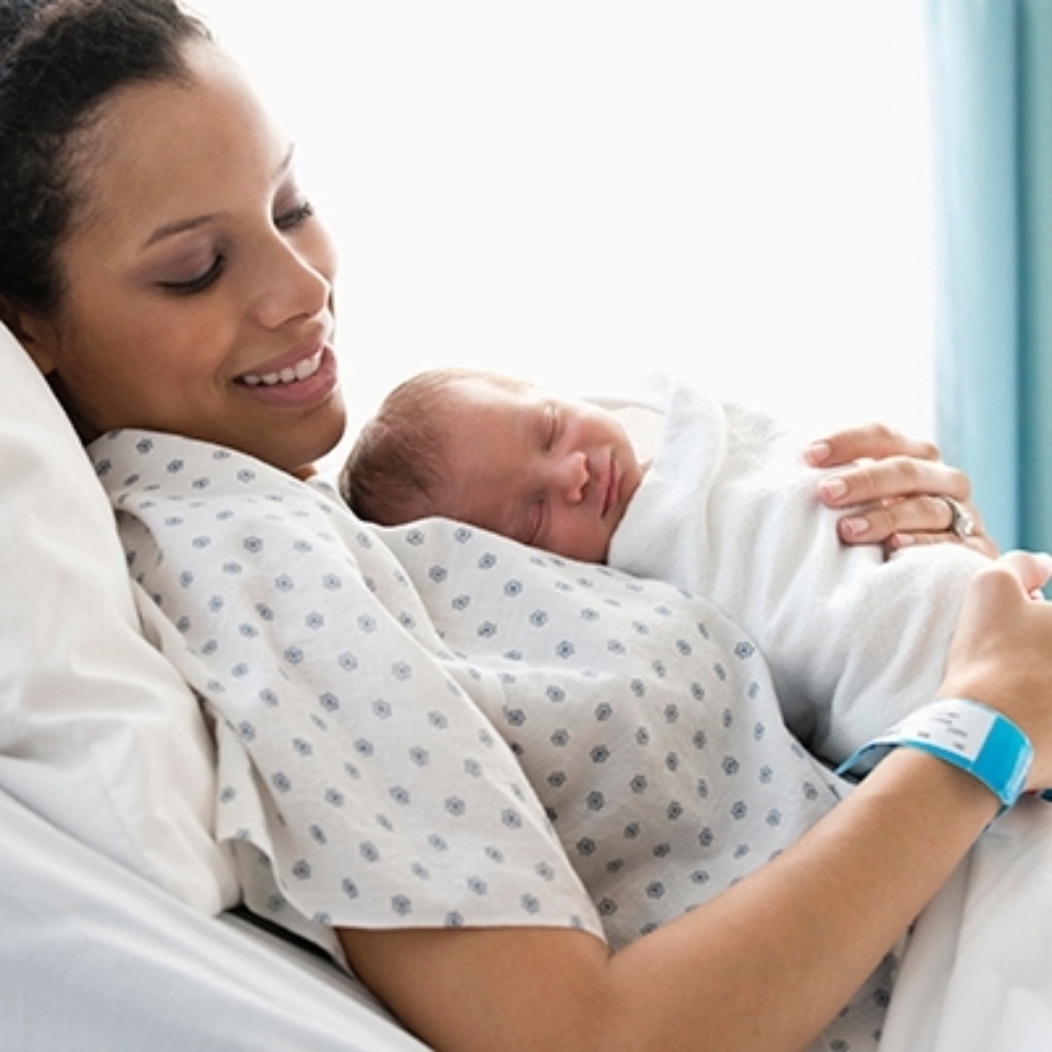 Smiling mother in a hospital gown sits with her newborn baby resting on her chest