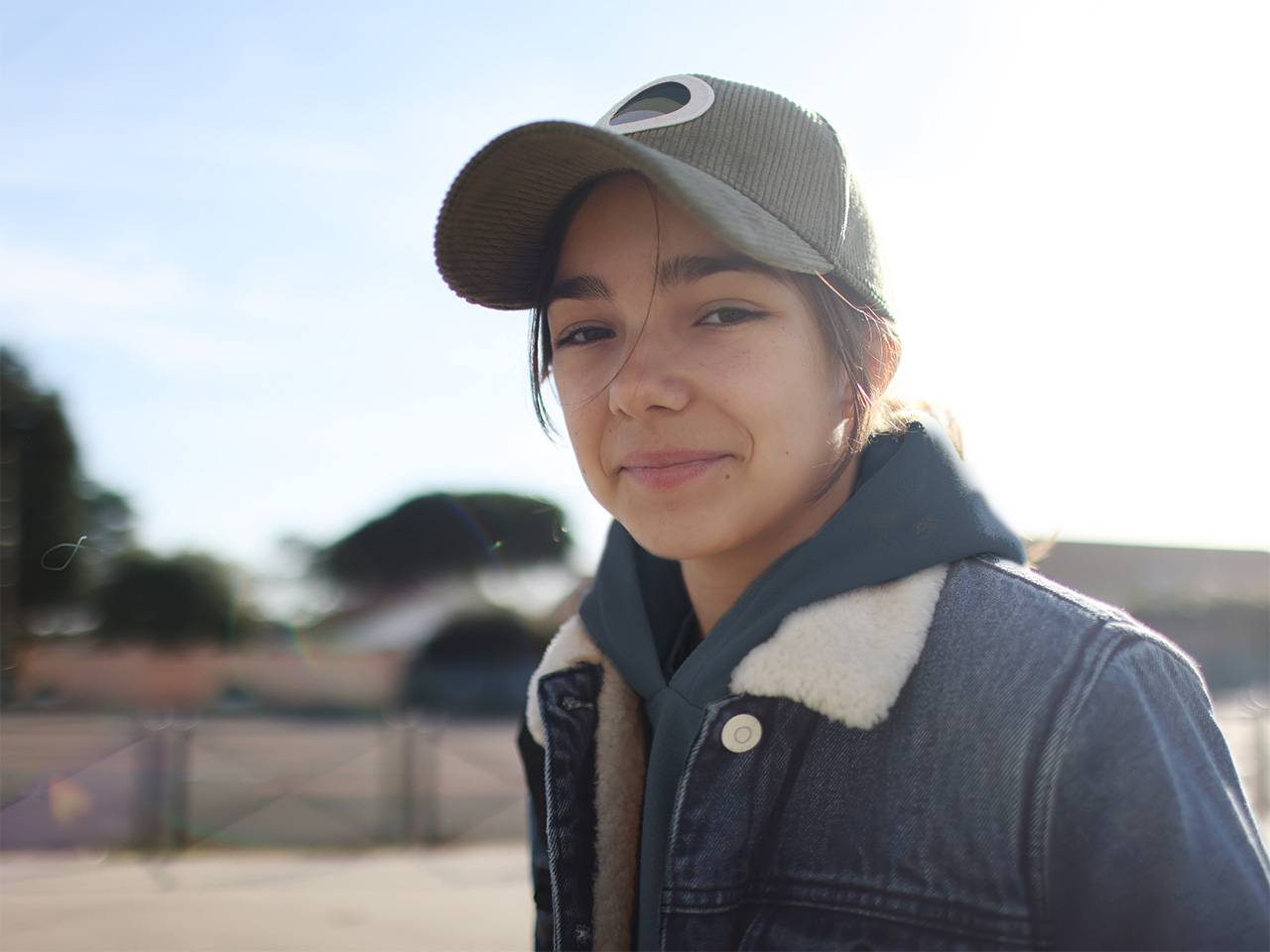 A teenager wearing a denim jacket and a ballcap smiles as they stand outdoors in the sunshine