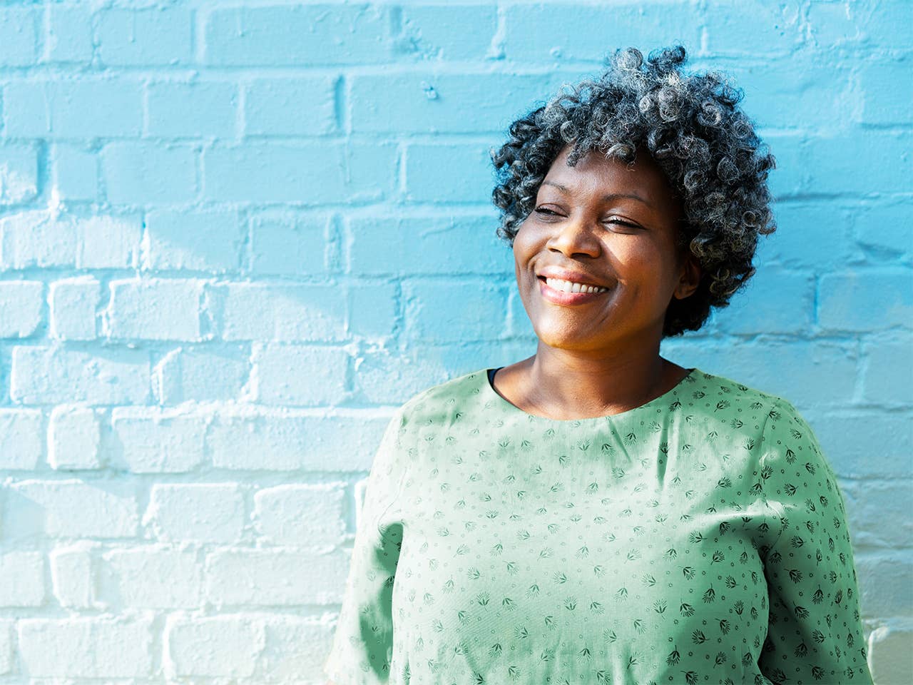 Smiling middle aged Black woman wearing a light green blouse stands outside in front of a white brick wall